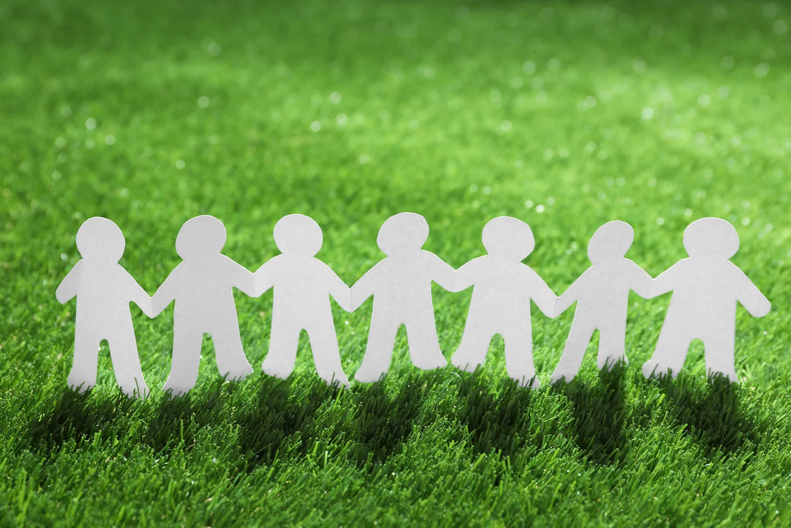 Paper figures holding hands on green grass, representing teamwork, community unity, collaboration, and social responsibility.