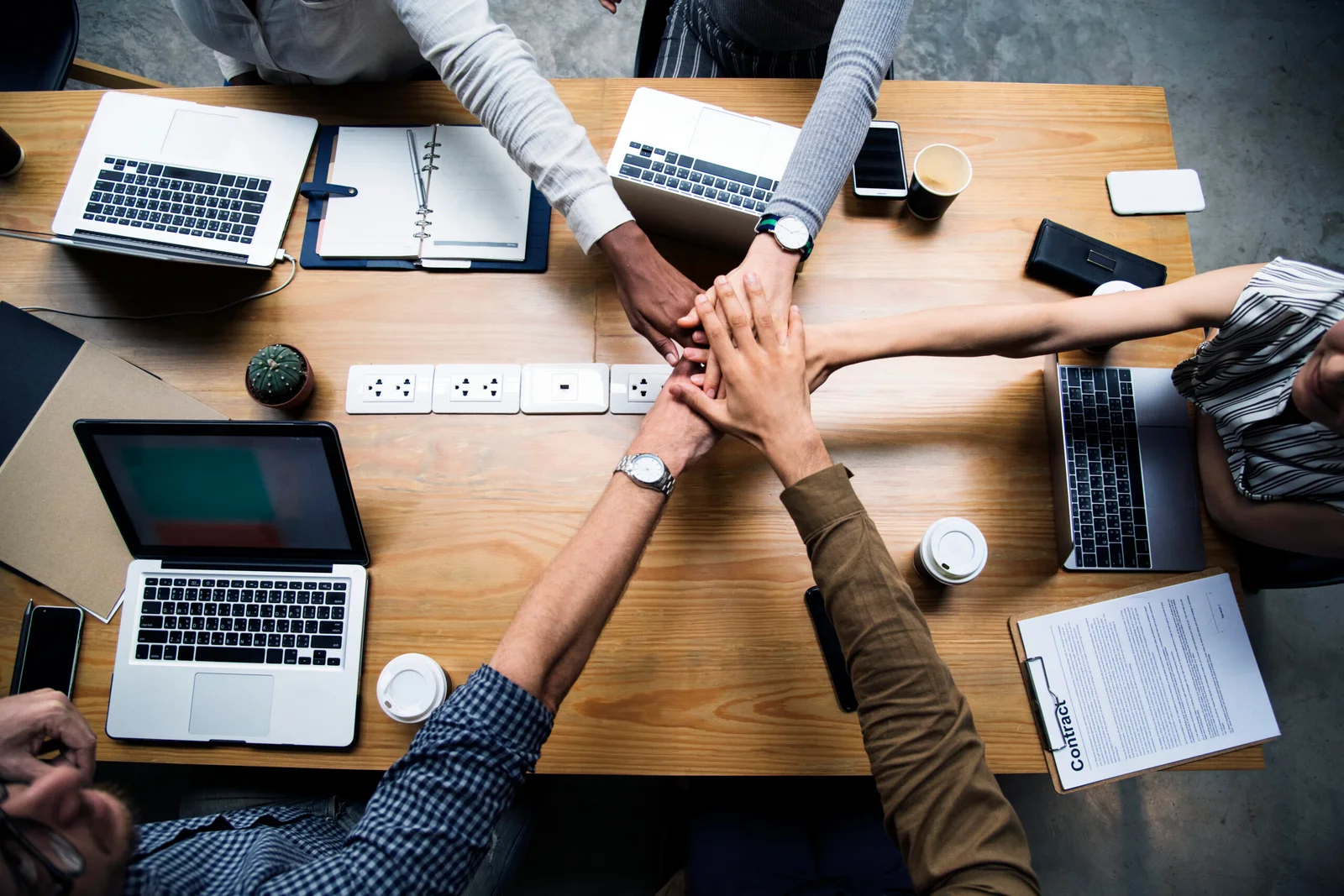 Team members joining hands over a conference table with laptops and documents, representing collaboration and teamwork.